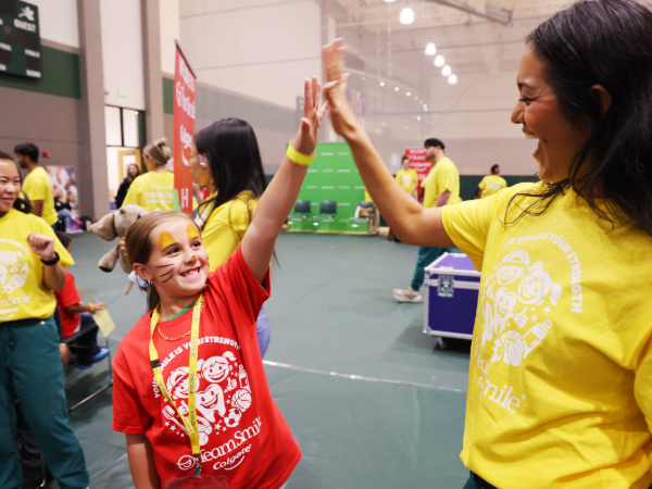 A smiling girl with face-painted whiskers in a red TeamSmile t-shirt high-fives a dental student volunteer wearing a yellow TeamSmile shirt at a lively community event in a gymnasium.